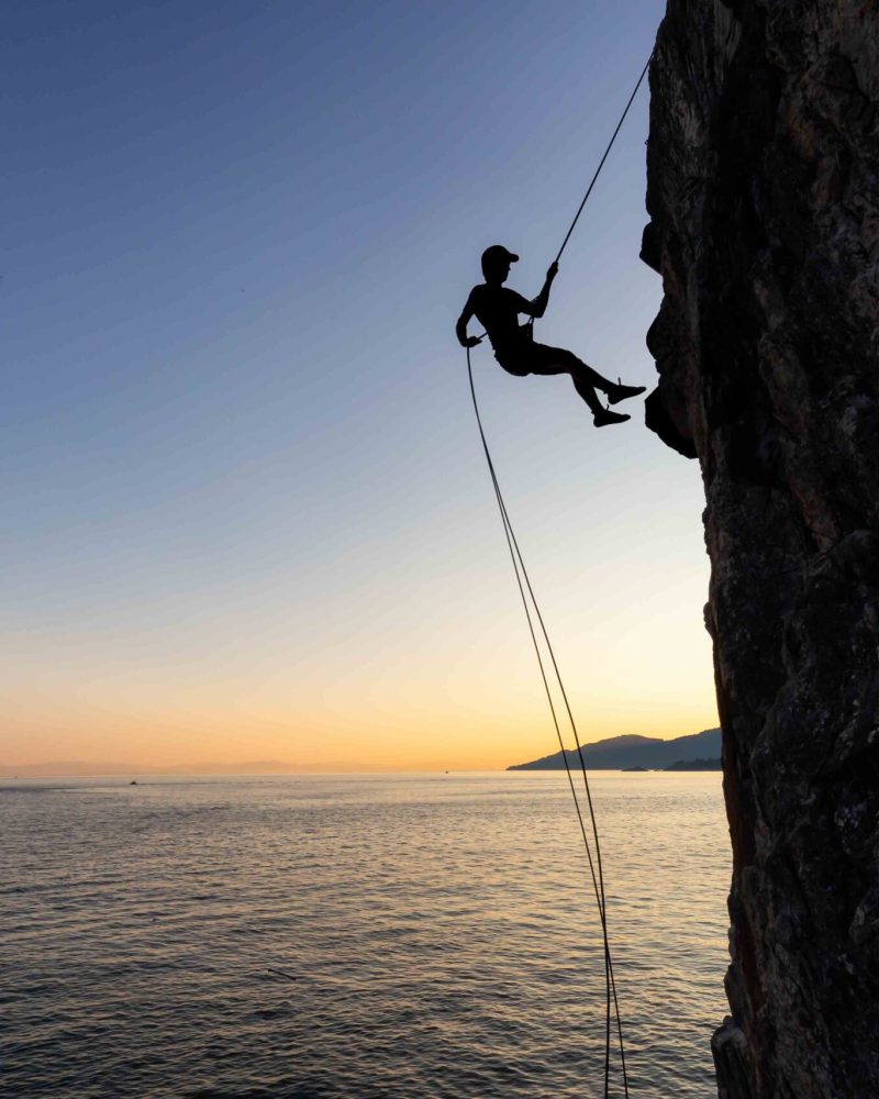 Silhouette of a Unrecognizable man rappelling down a steep cliff on the rocly ocean coast during a sunny summer sunset. Taken in Lighthouse Park, West Vancouver, British Columbia, Canada.