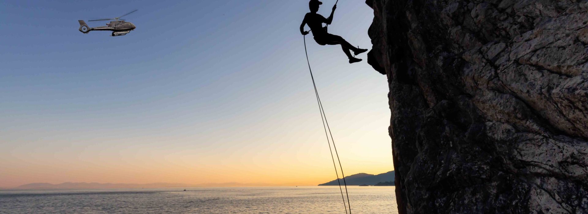 Silhouette of a Unrecognizable man rappelling down a steep cliff on the rocly ocean coast during a sunny summer sunset. Taken in Lighthouse Park, West Vancouver, British Columbia, Canada.