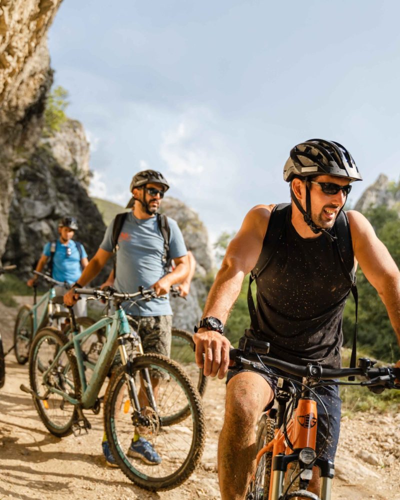 man caucasian male cyclist riding e-bike electric bicycle outdoor in mountain range at the gorge or ridge in front of his friends wear protective helmet and eyeglasses in sunny day copy space