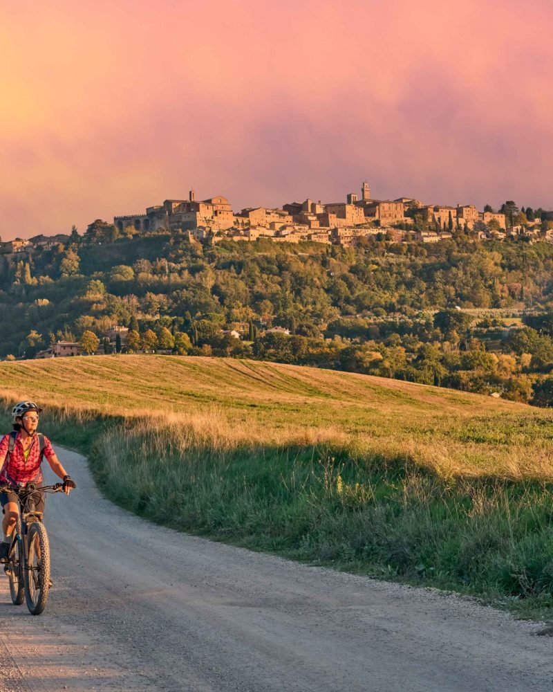 nice senior woman riding her electric mountain bike between olive trees in the Ghianti area with Montepulciano in background, Tuscany , Italy