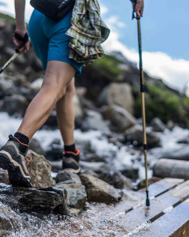 Woman wearing outdoor boots and shorts walking across a wooden bench and rocks crossing a wild alpine creek using hiking poles. Trekking shoes on rocks and in runnig water.