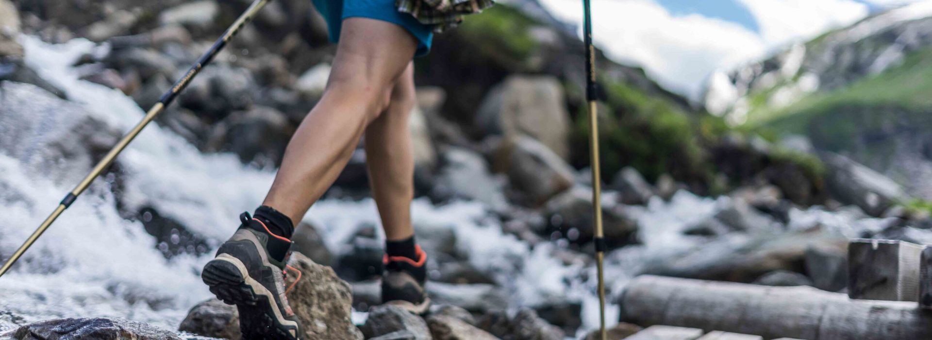 Woman wearing outdoor boots and shorts walking across a wooden bench and rocks crossing a wild alpine creek using hiking poles. Trekking shoes on rocks and in runnig water.